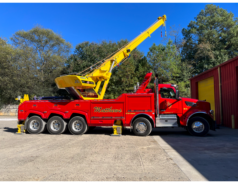 Red heavy-duty tow truck with yellow crane boom extended, parked beside a red building with a yellow door under blue sky