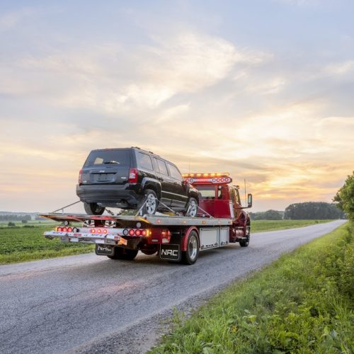 Tow truck carrying a black SUV on a rural road at sunset, with flashing amber lights and open fields on both sides