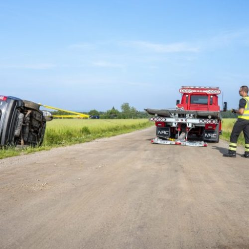 Tow truck on dirt road preparing to recover an overturned SUV in a grassy ditch, worker in reflective vest nearby