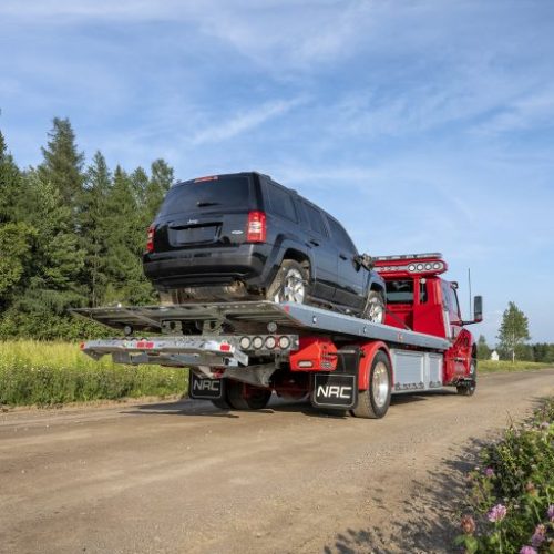 Red flatbed tow truck hauling a black SUV on a rural dirt road beside wildflowers under a blue sky