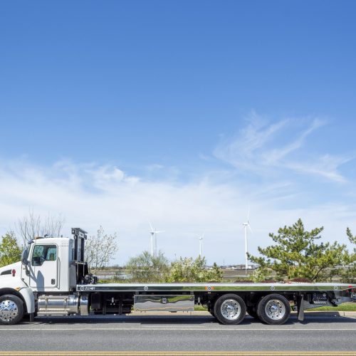 Side view of a white flatbed tow truck parked on a road with wind turbines and trees under a clear blue sky