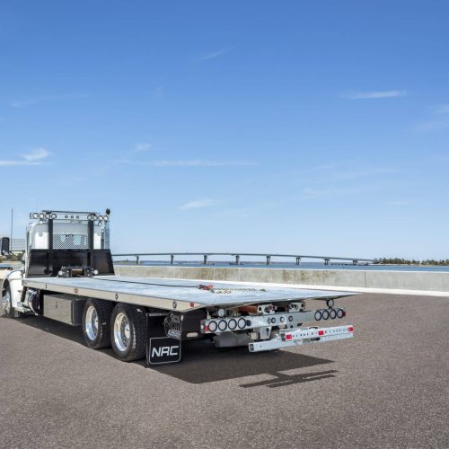Rear view of a flatbed tow truck on a roadway beside water, with city skyline and bridge in the background