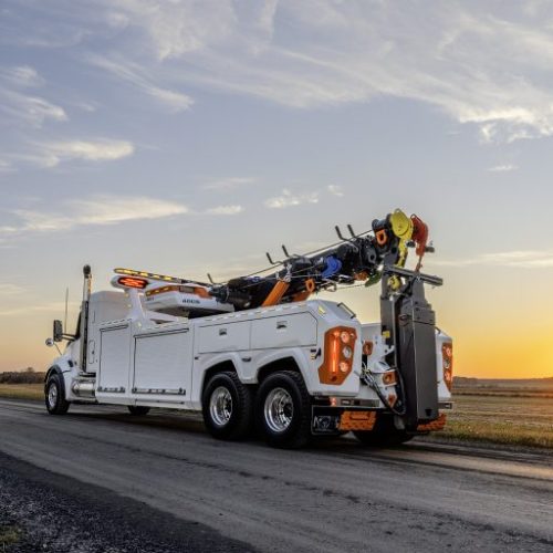 White heavy-duty tow truck on a rural road at sunset, with recovery boom and winches mounted on the back