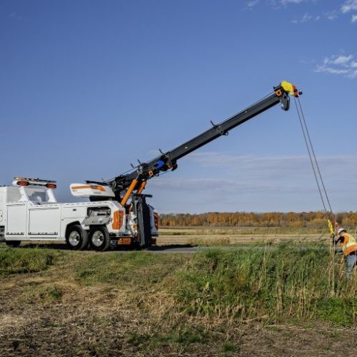 White utility truck with boom crane lifting cables as a worker in a safety vest guides the line in a grassy field under blue sky