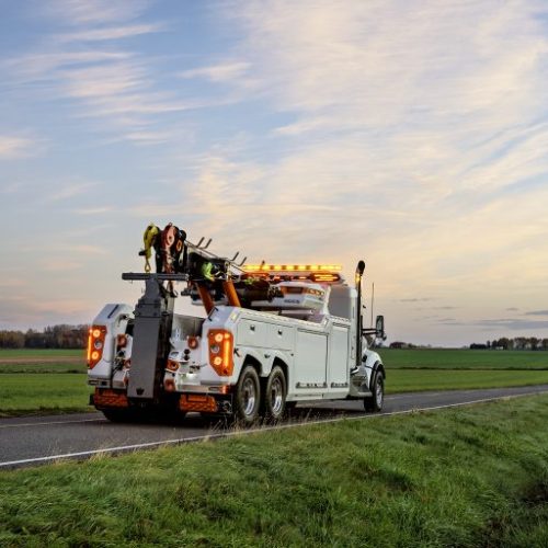 Tow truck with flashing amber lights parked on a rural road at dusk, with open fields and a pastel sky in background