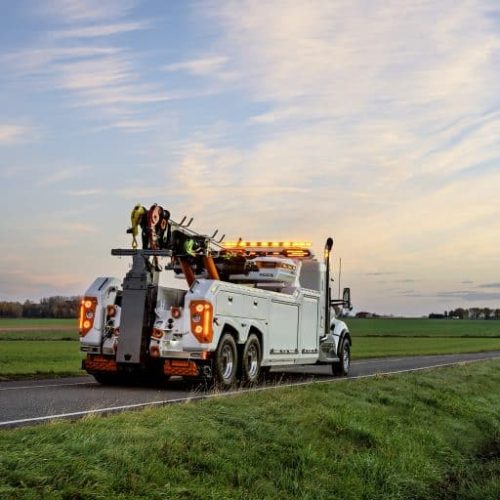 Tow truck with flashing amber lights parked on a rural road at dusk, with open fields and a pastel sky in background
