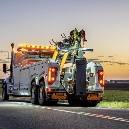 Heavy-duty tow truck with amber lights on a rural highway at dusk, viewed from the rear with flat fields in the background
