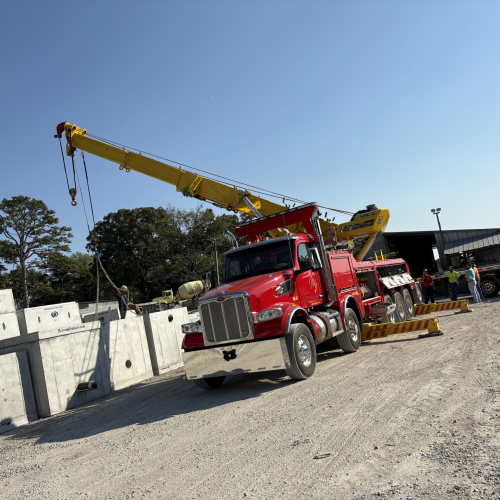 Red crane truck lifting precast concrete sections at a construction yard, with workers nearby under a clear blue sky