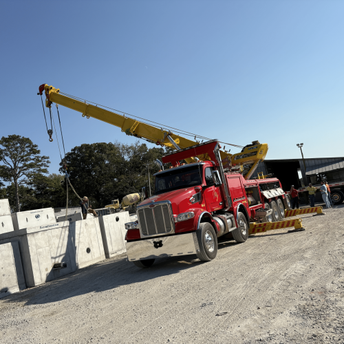 Red crane truck lifting precast concrete sections at a construction yard, with workers nearby under a clear blue sky