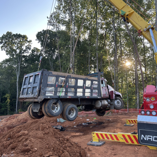 Crane lifting a dump truck at a construction site with dirt piles and trees in the background at sunset