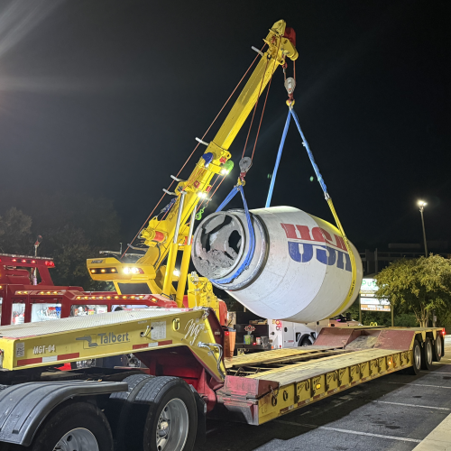 Yellow crane hoisting a large concrete mixer drum onto a lowboy trailer at night under bright parking lot lights