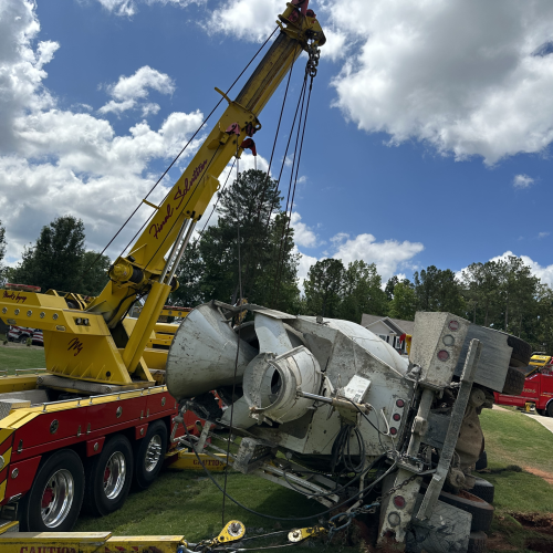 Yellow tow crane lifting an overturned cement mixer truck on a grassy yard under a blue sky with clouds