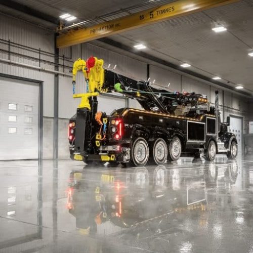 Black heavy-duty tow truck with yellow crane boom parked inside a large industrial garage with glossy wet floor reflections