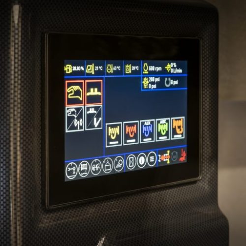 Touchscreen control panel on industrial machine displaying system icons and operating data in a dark carbon-fiber housing