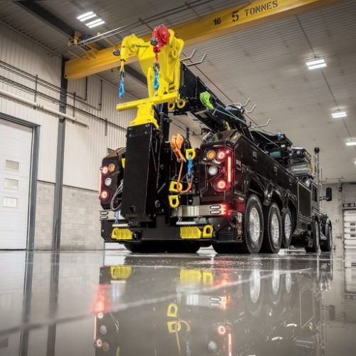 Heavy-duty tow truck with yellow boom and rigging inside a large industrial garage with glossy floor reflections