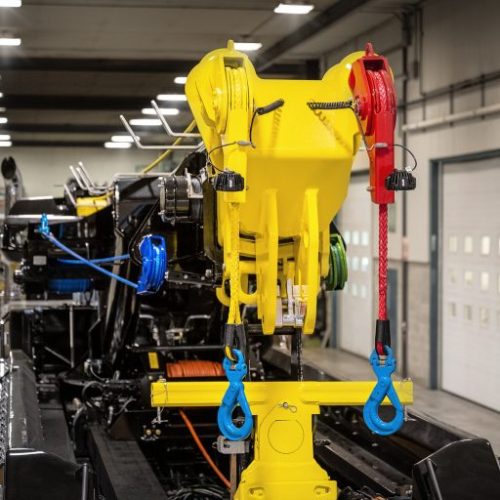 Yellow and red industrial hoist with blue hooks mounted on machinery inside a large workshop or factory floor