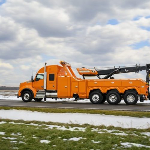 Orange heavy-duty tow truck driving on a rural road beside snow-covered fields under a cloudy sky