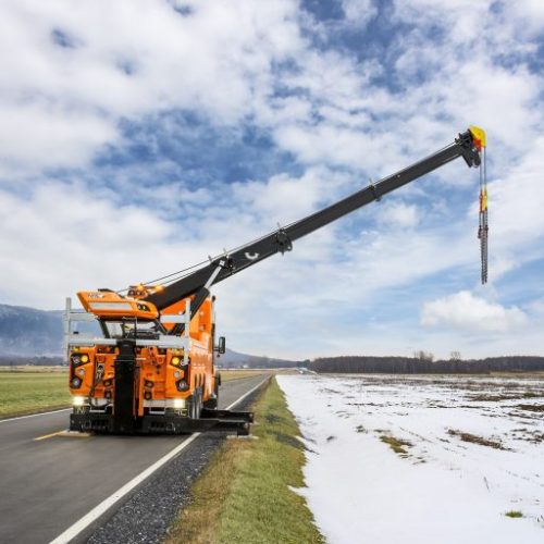 Orange crane truck with extended boom and hanging chains parked on rural road beside snowy field under cloudy sky