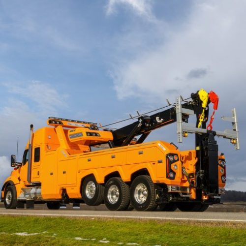Orange heavy-duty tow truck with boom and rigging parked on a roadside under a cloudy sky