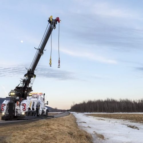 Heavy-duty tow truck with extended boom and hanging hooks on a rural road at dusk beside snowy fields