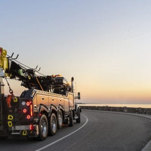 Heavy-duty tow truck driving on a coastal road at sunset, with recovery boom and equipment on the back