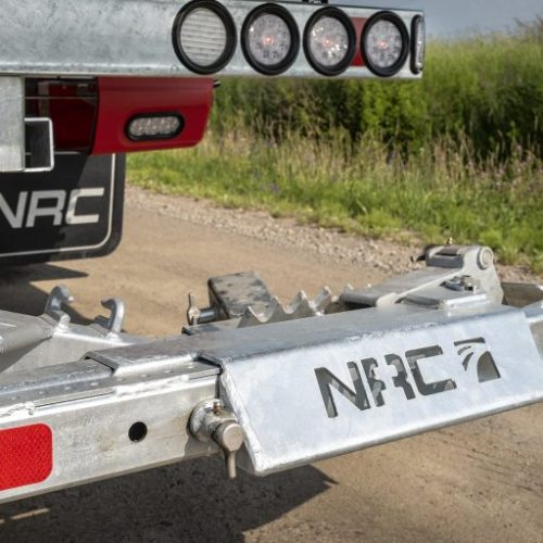 Close-up of a galvanized trailer hitch and metal accessory plate with NRC logo, parked on a gravel road beside grass