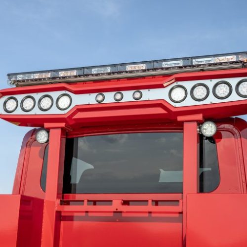 Close-up of red emergency vehicle cab with roof-mounted LED light bar and multiple round spotlights against blue sky