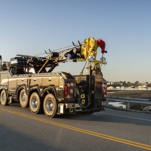 Heavy-duty tow truck parked on a coastal road at sunset, with harbor and boats in the background
