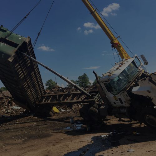 Crane lifting a large metal container beside a damaged truck in a scrapyard under a blue sky