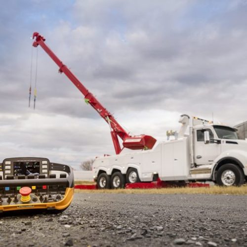 Remote control unit in foreground with white tow truck and red boom crane in background under cloudy sky