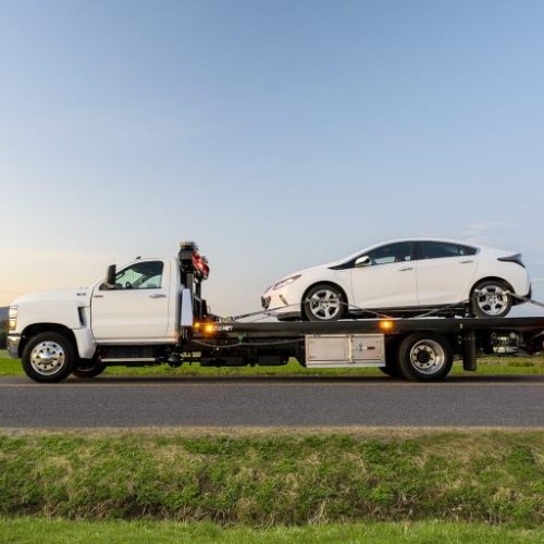 Flatbed tow truck carrying a white sedan on a rural road at sunset