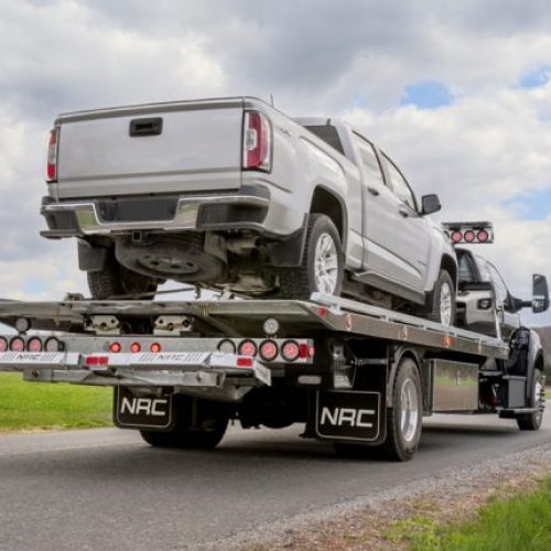 Car carrier flatbed truck hauling a silver pickup on a rural road under cloudy sky