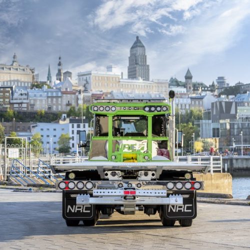 Rear view of green flatbed tow truck on waterfront quay with city skyline and cloudy sky in the background