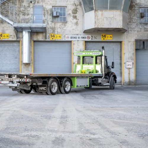 Green and black flatbed tow truck parked in front of industrial building with multiple roll-up garage doors