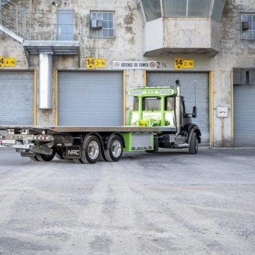 Green and black flatbed tow truck parked in front of industrial building with multiple roll-up garage doors