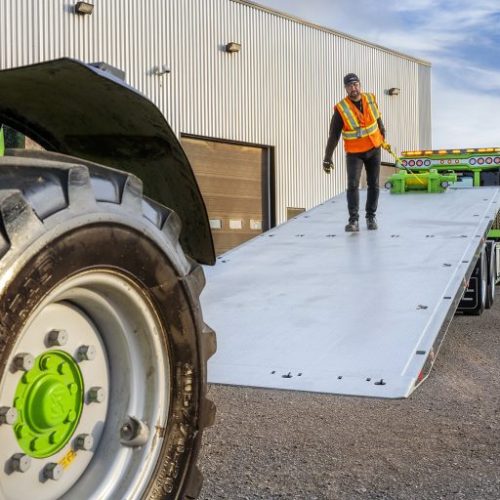 Worker in safety vest on a long metal loading ramp beside a large tractor tire outside an industrial building