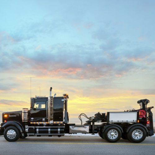 Side view of black heavy-duty rollback tow truck on a roadside by the water at sunset with pastel clouds in the sky
