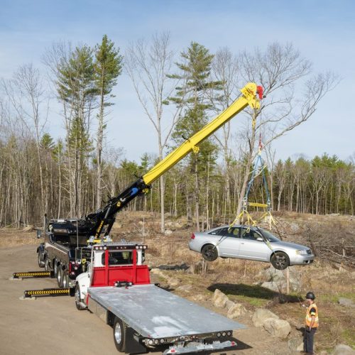 Tow truck with crane lifts a silver sedan off rocky roadside in a wooded clearing while a worker in safety vest watches