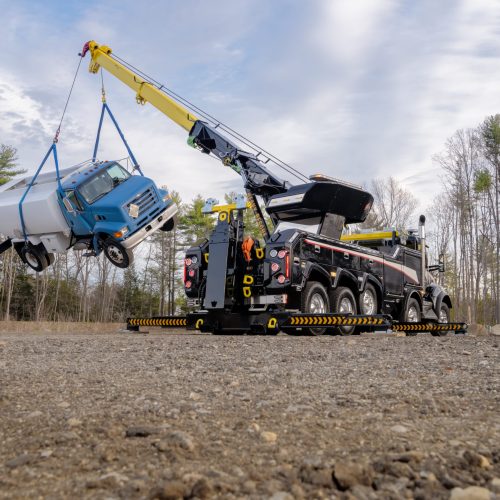 Heavy-duty recovery tow truck crane lifting a blue tanker truck with straps in a gravel lot beside trees under cloudy sky